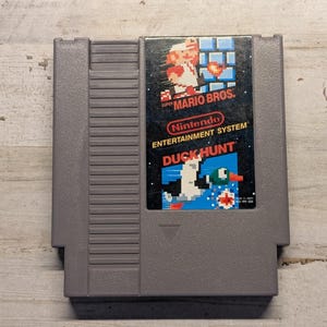 May include: A gray Nintendo Entertainment System game cartridge for Super Mario Bros. and Duck Hunt. The label features pixelated graphics of Mario and a duck, with the text Super Mario Bros., Nintendo Entertainment System, and Duck Hunt printed.