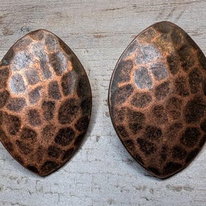 May include: A pair of oval-shaped earrings with a hammered copper finish. The metal surface features a dark, mottled patina with raised, rounded textures that create a distinct pattern across the front of each piece, showcasing a metallic sheen.