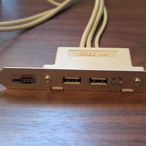 May include: An MSI branded computer expansion bracket featuring two USB ports on a silver metal faceplate. The component includes a beige plastic housing and attached cables, with a visible USB logo and four small circular indentations on the front.