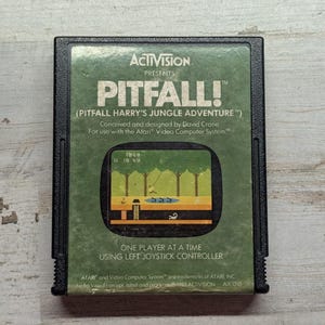 May include: A black rectangular Atari video game cartridge for Pitfall! featuring a green label with white text. The label displays the title, game credits, and a central graphic showing a pixelated jungle scene with a character swinging on a vine.