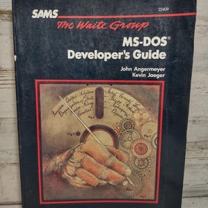 May include: A black paperback book titled MS-DOS Developer's Guide by The Waite Group, John Angermeyer, and Kevin Jaeger. The cover features a central illustration of a hand holding a stylus over a floppy disk graphic with gears and text.