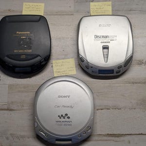 May include: Three portable CD players including a black Panasonic model with XBS branding, a silver Sony Discman ESP2 with Groove technology, and a silver Sony Walkman Car Ready player with ESPMAX, each featuring various control buttons and displays.