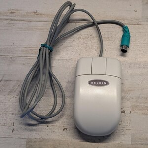May include: A light gray Belkin computer mouse featuring two primary buttons and a central scroll wheel. The device is connected to a long gray cord that terminates in a teal-colored circular PS/2 connector plug for legacy computer hardware.