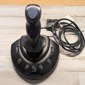May include: A black InterAct Raider Pro joystick featuring a central handle with a grey top button, a curved base with four grey circular buttons, two side-mounted scroll wheels, and a black coiled cable with a 15-pin game port connector attached.