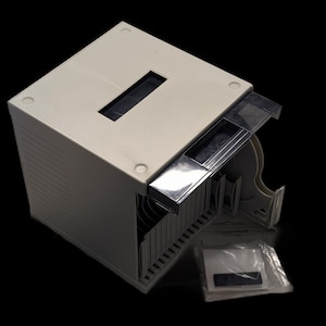 May include: A beige plastic storage case for 3.5-inch floppy disks featuring a rectangular top slot, a hinged transparent lid, and internal dividers. A small plastic bag containing a white label and a black adhesive strip is included.