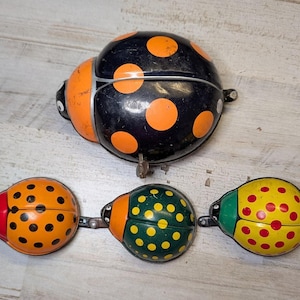 May include: A collection of four metal ladybug-shaped bicycle bells. One large black bell with orange spots, and three smaller bells featuring orange with black spots, green with yellow spots, and yellow with red spots, each with a metal lever.