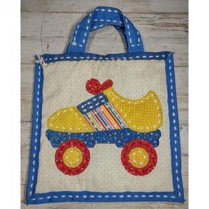 May include: A square fabric tote bag with a cream-colored background featuring a small brown polka dot pattern. The bag has a blue border and handles. A large applique of a roller skate in yellow, red, and striped fabric is centered.
