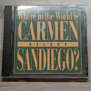 May include: A square CD jewel case featuring a dark green cover with tan text that reads Where in the World is CARMEN SANDIEGO? DELUXE. The title is displayed in large, bold, serif lettering centered on the front of the plastic case.