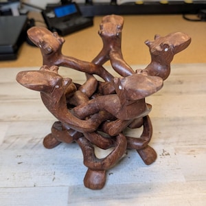 May include: A brown wooden display stand carved from a single piece of wood, featuring five interconnected figures with small white dot eyes. The structure forms a circular base and a top ring designed to hold an object, with a smooth finish.