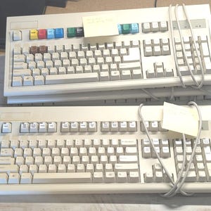 May include: Two beige computer keyboards with grey cables. The top keyboard features a row of multi-colored function keys including yellow, red, blue, green, black, and teal. Both keyboards have standard QWERTY layouts and numeric keypads.