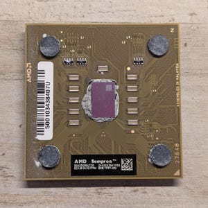 May include: A square AMD Sempron processor with a tan circuit board base, featuring a central purple die surrounded by silver thermal paste. It includes a white barcode label, four dark circular pads, and text reading AMD Sempron SDA25000UT3D.