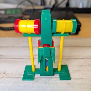 May include: A plastic toy oil derrick model featuring a green frame, yellow support poles, and two horizontal yellow barrels with red and green end caps. The structure includes a central vertical component and a small blue marble visible inside.