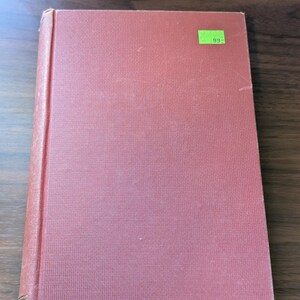 May include: A hardcover book with a solid red fabric cover and a textured surface. The book features a small, rectangular, bright green sticker on the upper right corner with the number 99 printed in black text. The spine is visible on the left.