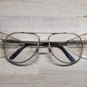 May include: Aviator-style glasses featuring a thin silver-toned metal frame and clear lenses. The design includes prominent, angular, translucent side shields attached to the outer edges of the frame, extending outward from the main lens area.