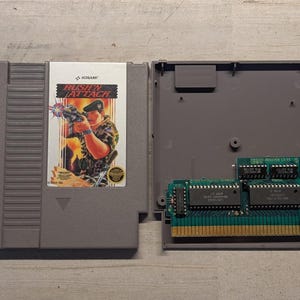 May include: A grey Nintendo Entertainment System game cartridge for Rush'n Attack, shown with its front label featuring a soldier and the internal green circuit board exposed on the right side, revealing the gold connector pins and black microchips.