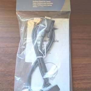 May include: A grey Palm HotSync Cable for III and VII series devices, sealed in a clear plastic retail package. The packaging features the Palm logo, the text HotSync Cable, and includes a white instruction manual visible behind the coiled cable.