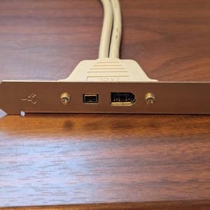 May include: A beige computer expansion card bracket featuring a metallic silver faceplate with two ports, including one small rectangular port and one larger rectangular port, secured by two screws, with two beige cables extending from the top.