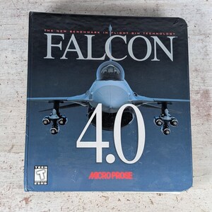 May include: A Falcon 4.0 flight simulator video game box featuring a front-facing fighter jet graphic, the title text in white, the MicroProse logo in red, and a Teen T ESRB rating symbol in the bottom left corner on a dark blue background.