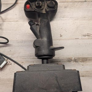 May include: A black flight joystick controller featuring a textured grip, a prominent red button on the top left, a circular directional hat switch on the top right, and a secondary trigger button on the side, mounted on a rectangular base.