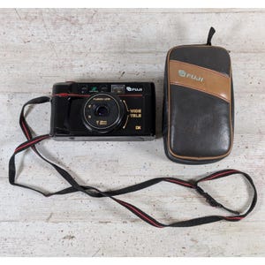 May include: A black Fuji film camera with a Fujinon lens, featuring Wide Tele and DX markings, accompanied by a black and brown Fuji branded carrying case. A black camera strap with a thin red stripe is attached to the camera body for portability.