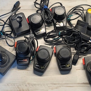 May include: A collection of black video game controllers including two joysticks with red buttons and several paddle controllers with circular knobs and black cords. Each controller features a label with text such as paddle or driving.