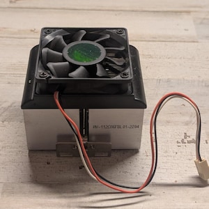 May include: A black computer cooling fan mounted on a silver metal heatsink. The fan has a circular green center and black blades. A red, black, and white wire extends from the fan to a white connector. The text AV-112C86FBL 01-2204 is printed on the side.