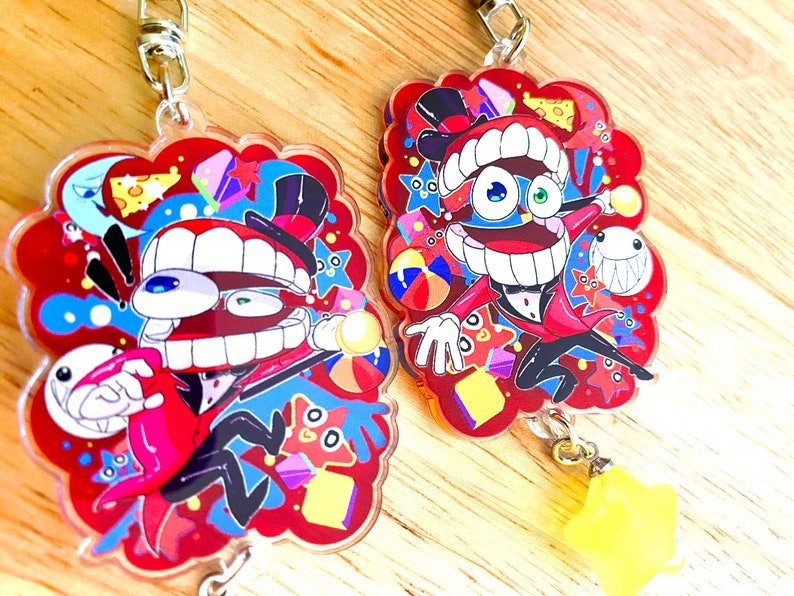 May include: Two acrylic keychains featuring cartoon characters with large mouths, top hats, and red suits. The keychains have a red background with colorful shapes and a yellow star charm. Each keychain is approximately 3 inches in height.