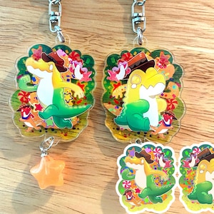 May include: Two clear acrylic keychains featuring a cartoon alligator wearing a cowboy hat. The alligator is yellow and green, with a star-shaped charm hanging from one keychain. The background is decorated with colorful candies and other cartoon characters.