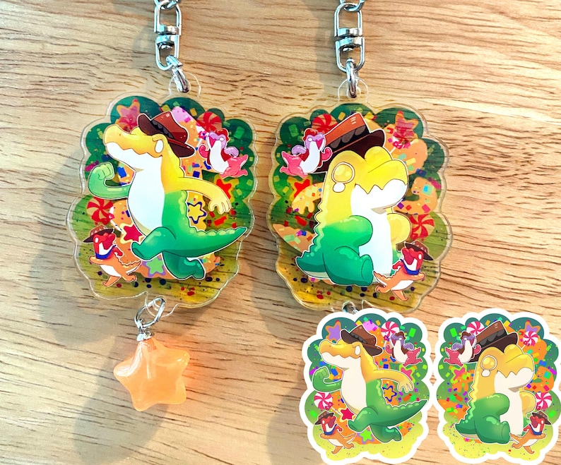May include: Two clear acrylic keychains featuring a cartoon alligator wearing a cowboy hat. The alligator is yellow and green, with a star-shaped charm hanging from one keychain. The background is decorated with colorful candies and other cartoon characters.