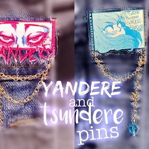 May include: Two enamel pins on a denim background. The left pin is pink and features a pair of red eyes with the word "Yandere" written in pink and white. The right pin is blue and features a cartoon girl with the word "Tsundere" written in blue and white.
