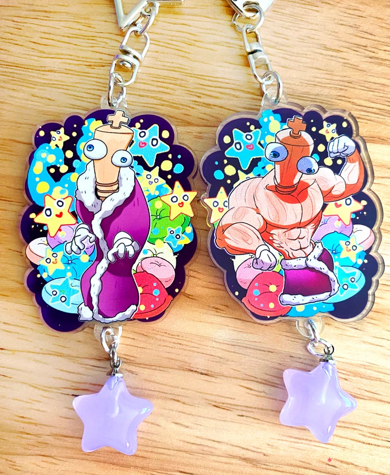 May include: Two cartoon keychains featuring a kingly character and a muscular character. The kingly character wears a purple robe with a white fur trim. The muscular character is shirtless. Each keychain has a star charm.