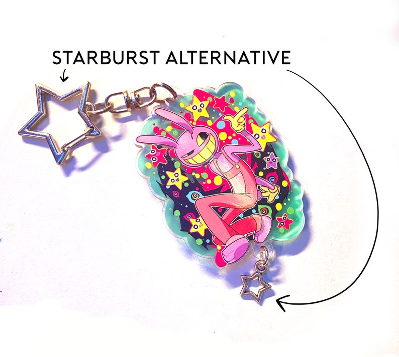 May include: A keychain featuring a cartoon character with a pink bunny-like appearance, wearing a pink jumpsuit, and a wide grin. The character is set against a colorful background of stars and shapes. The keychain has a star-shaped clasp and a small star charm.