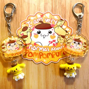 May include: Two yellow and white Pompompurin character keychains with a yellow and white Pompompurin character sticker. The sticker says "I am very normal about Pompompurin".