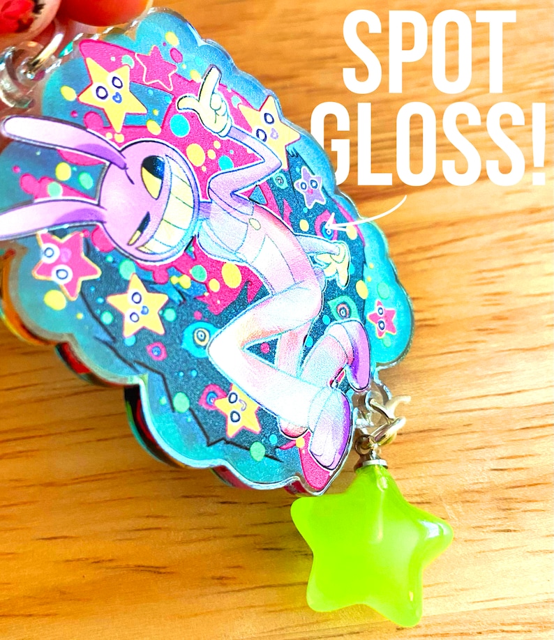 May include: A colorful keychain featuring a cartoon character with a large grin, surrounded by stars and a blue cloud-like shape. The keychain has a neon green star charm and the text "SPOT GLOSS!"