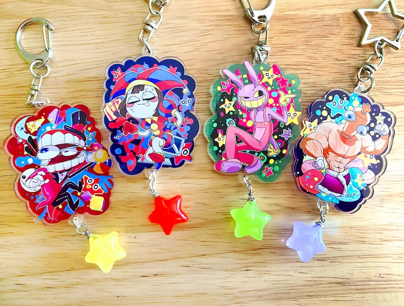 May include: Four colorful acrylic keychains with cartoon character designs. Each keychain features a unique character illustration with a star-shaped charm. The keychains have silver-colored hardware and are displayed on a wooden surface.