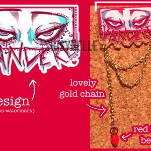 May include: A 1.5 inch acrylic pin with a design of two eyes and the word "VANDEREY" in pink, blue, and white. The pin is attached to a gold chain with a red teardrop bead.
