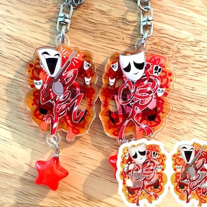 May include: Two keychains with a theatrical theme. Each keychain features a red and white figure with a mask, holding a musical instrument. The background is orange with red accents. One keychain has a red star charm.