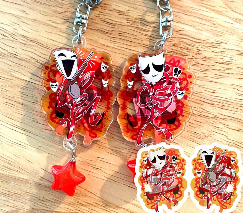 May include: Two keychains with a theatrical theme. Each keychain features a red and white figure with a mask, holding a musical instrument. The background is orange with red accents. One keychain has a red star charm.