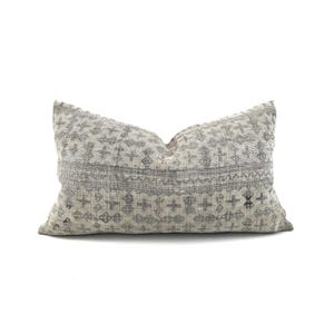 May include: Rectangular cushion with a grey and off-white geometric pattern. The cushion features a repeating cross and diamond design, with a horizontal band of detailed patterns. The cushion is made of a textured fabric.