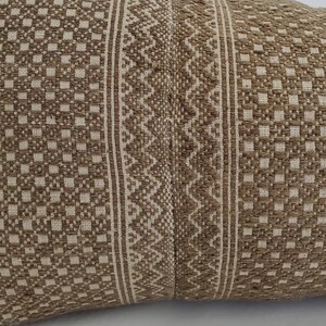 May include: Close-up of a woven pillow with a geometric pattern. The pillow features a repeating square pattern in a light beige color, with a central stripe of a zigzag design in the same beige color. The background is a darker brown.
