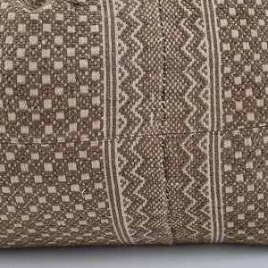 May include: Close-up of a woven pillow with a geometric pattern. The pillow features a repeating square pattern in a light beige color, with vertical stripes of a zigzag design in a darker brown color. The pillow is made of a textured fabric.