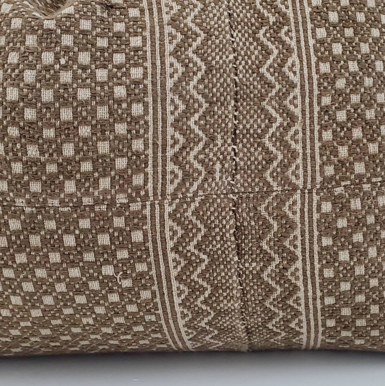 May include: Close-up of a woven pillow with a geometric pattern. The pillow features a repeating square pattern in a light beige color, with vertical stripes of a zigzag design in a darker brown color. The pillow is made of a textured fabric.