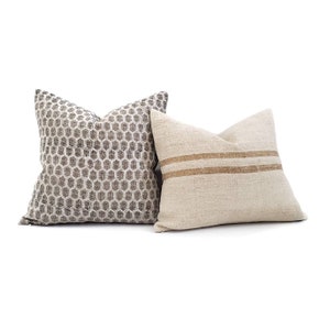 Puede incluir: Dos cojines decorativos. Uno presenta un patr&oacute;n geom&eacute;trico de panal en tonos grises y blancos. El otro es de color beige natural con dos rayas horizontales marrones. Ambos cojines son cuadrados y tienen un aspecto suave y texturizado.