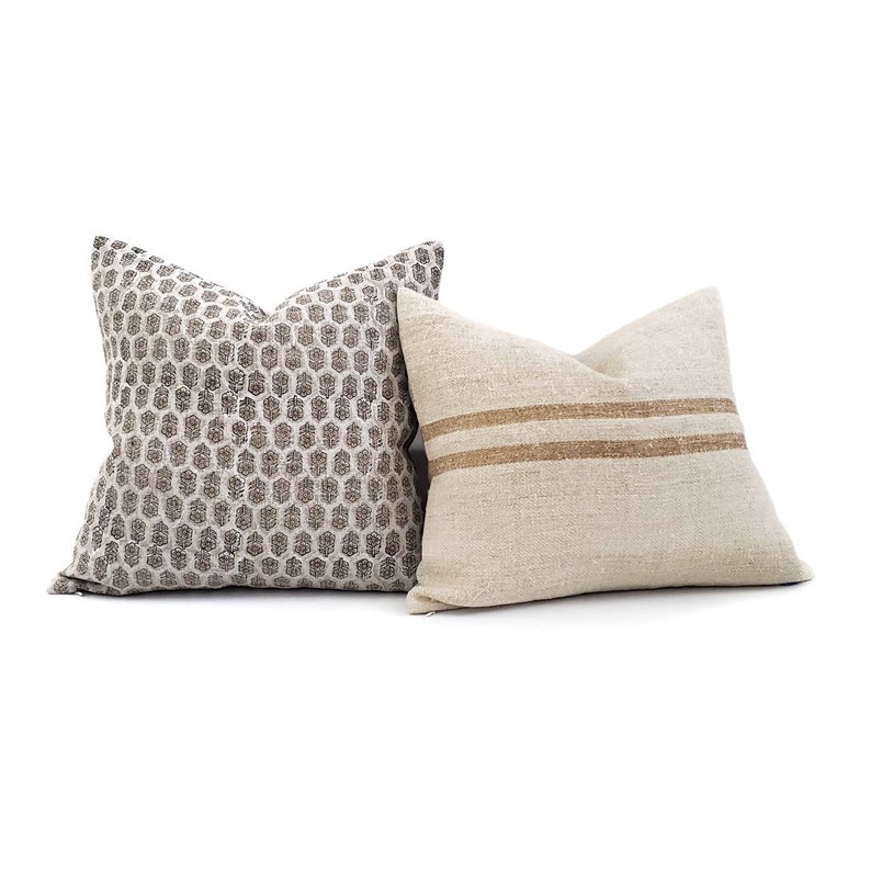 Puede incluir: Dos cojines decorativos. Uno presenta un patr&oacute;n geom&eacute;trico de panal en tonos grises y blancos. El otro es de color beige natural con dos rayas horizontales marrones. Ambos cojines son cuadrados y tienen un aspecto suave y texturizado.
