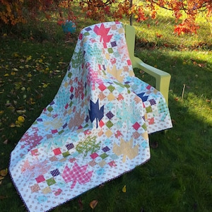 May include: A white quilt with a patchwork design featuring colorful leaves and squares. The quilt is folded and draped over a green chair in a grassy yard.