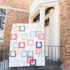 Peut inclure: Une courtepointe blanche avec un motif géométrique de carrés et de rectangles de différentes couleurs, dont le rouge, le bleu, le vert, le jaune et le rose. La courtepointe est accrochée à un mur de briques.