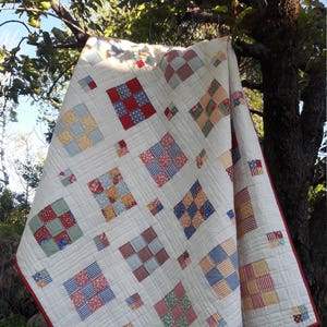Puede incluir: Un edredón hecho a mano con un fondo blanco y un diseño de patchwork. El edredón presenta cuadrados de varios tamaños y patrones en rojo, azul, amarillo y verde. Los bordes están acabados con un borde rojo. El edredón está colgado de un árbol.