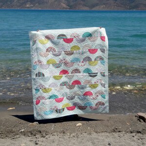 May include: A white quilt with a pattern of colorful half-circles. The quilt is displayed on a beach with a lake in the background.
