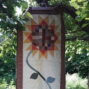 Può includere: Un quilt fatto a mano con un grande girasole con un centro a patchwork e uno stelo con due zucche. Il quilt è fatto con una varietà di tessuti in marrone, giallo, arancione e verde.