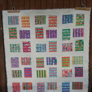 Peut inclure: Une courtepointe blanche avec un motif de patchwork composé de carrés colorés avec différents motifs rayés. La courtepointe est accrochée à un mur en bois avec une étiquette qui dit "Williams".